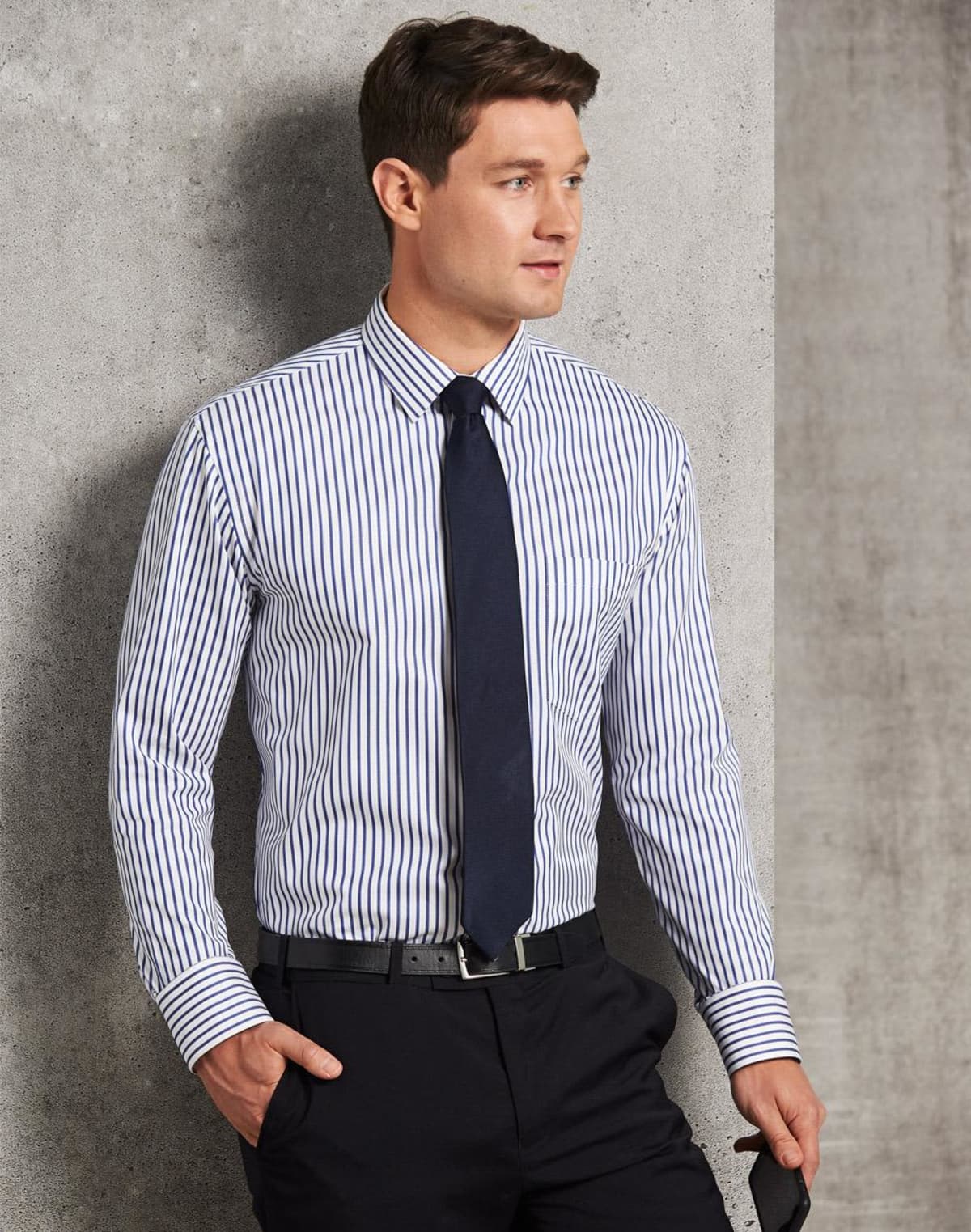Man wearing a striped dress shirt, black tie, and black pants standing against a concrete wall.