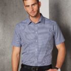 Man in a short-sleeve checkered shirt standing against a gray wall with one hand in his pocket.