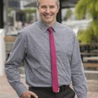 Smiling man in checked shirt and pink tie standing outdoors near benches and greenery.