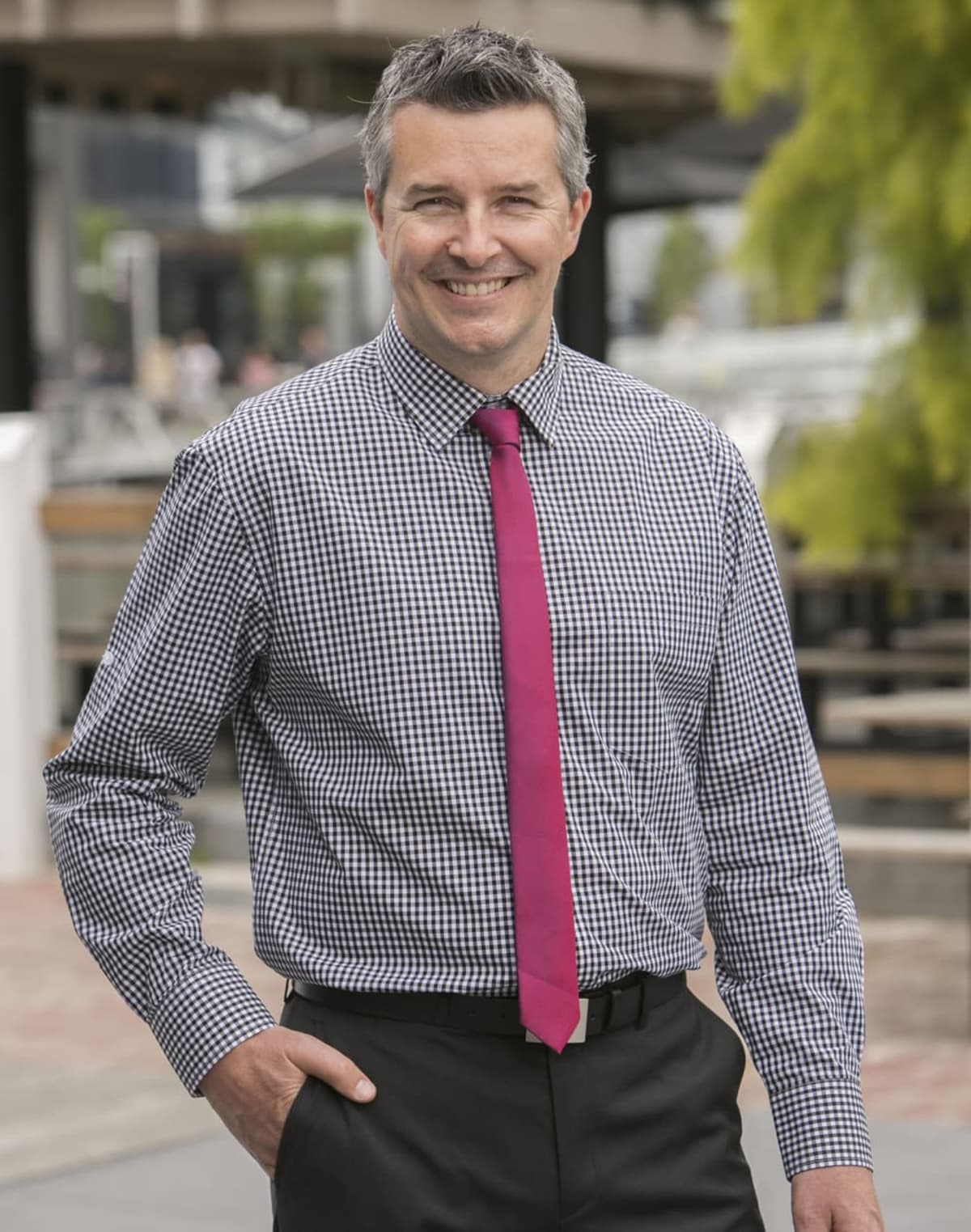 Smiling man in checked shirt and pink tie standing outdoors near benches and greenery.