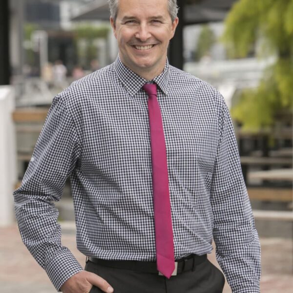 Man in a checkered shirt with a bright pink tie, smiling outdoors near benches and greenery.