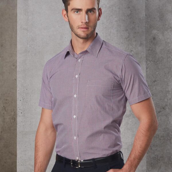Man in a short-sleeved checkered shirt and black pants standing against a gray wall.