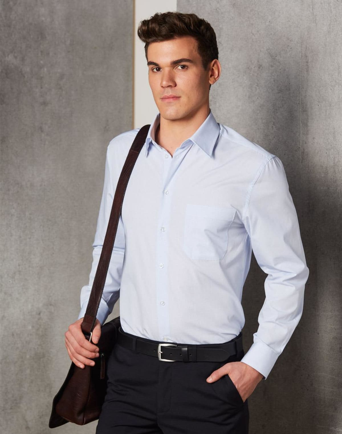Man in light blue dress shirt and black pants holding a shoulder bag, standing indoors.