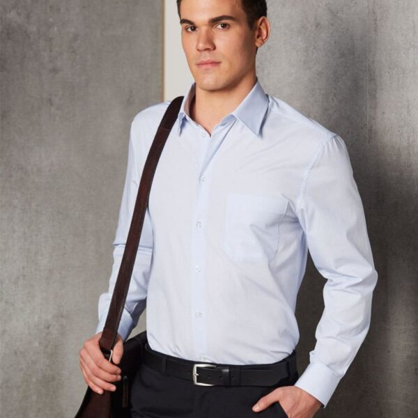 Man in a light blue dress shirt with a shoulder bag standing against a gray background.