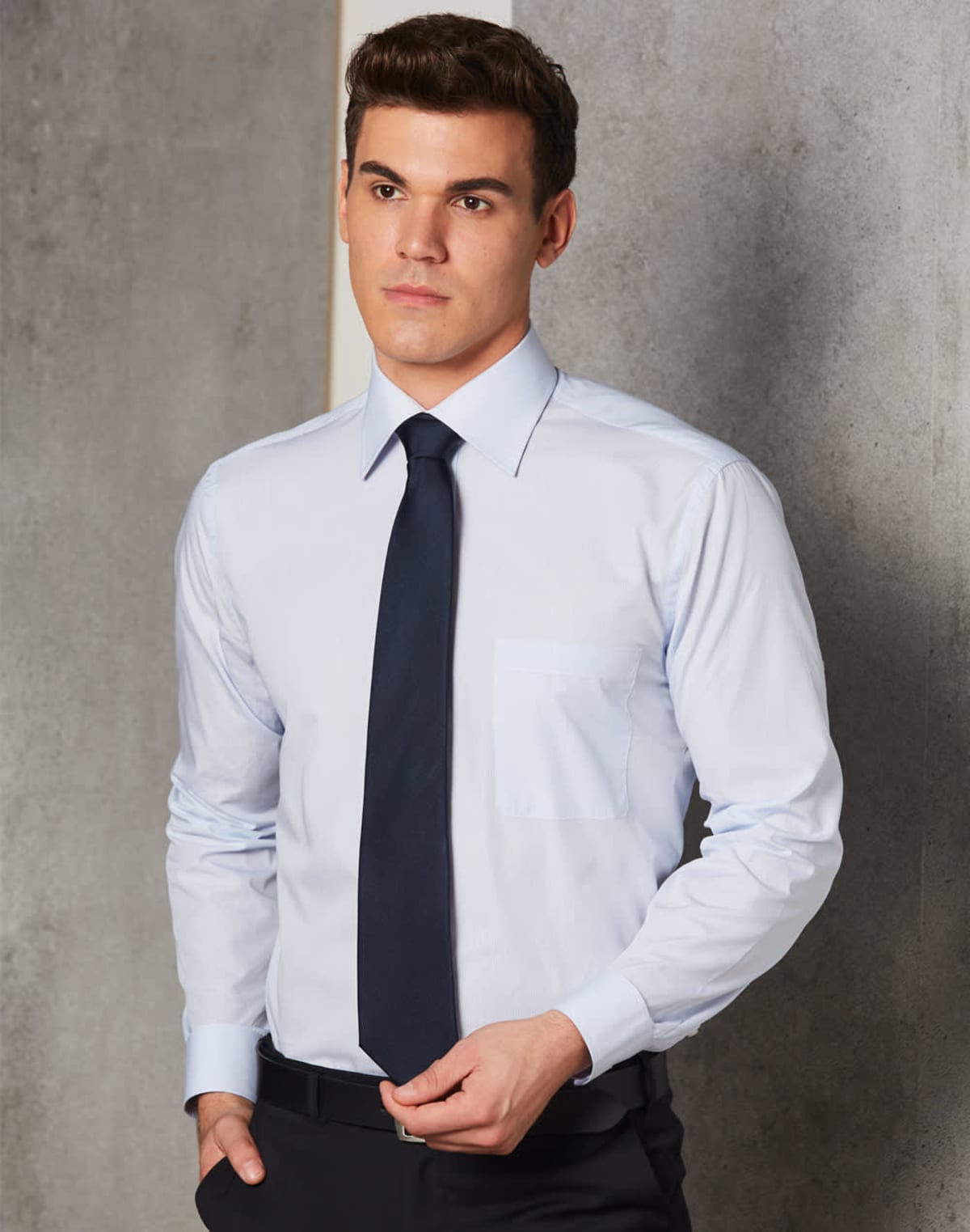 Man in a light blue dress shirt and dark tie standing against a gray wall.