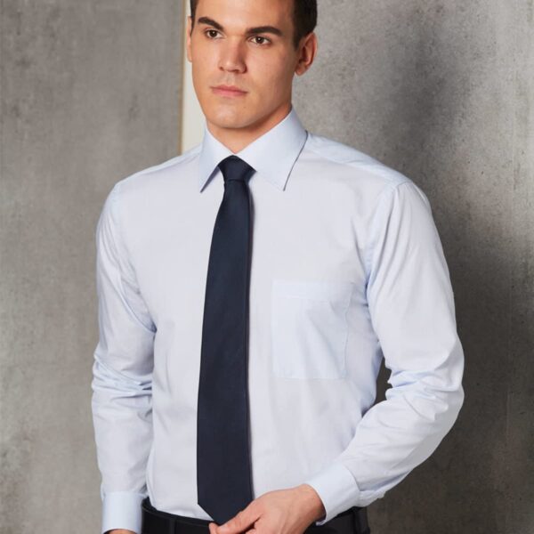 Man in a light blue dress shirt and dark tie standing against a gray wall.