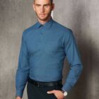 Man wearing a teal dress shirt and black trousers, standing against a gray textured wall.