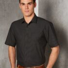Man in a black short-sleeve shirt and khaki pants standing against a textured wall.