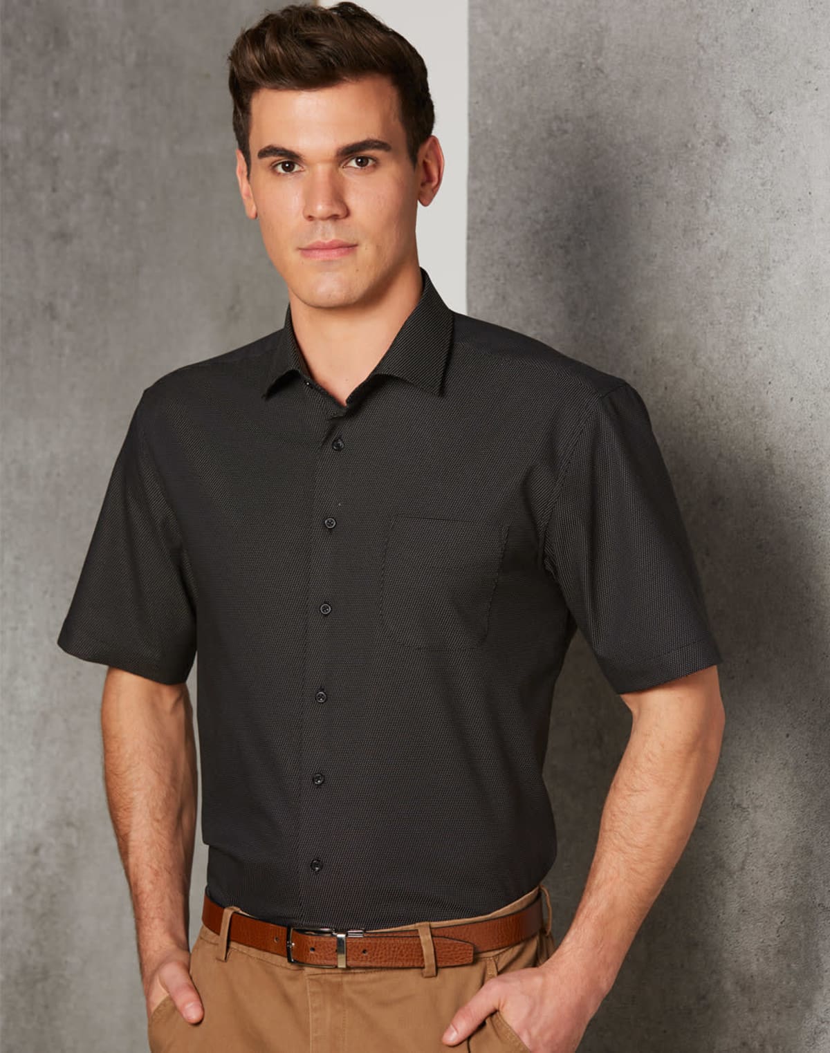 Man in a black short-sleeve shirt and khaki pants standing against a textured wall.