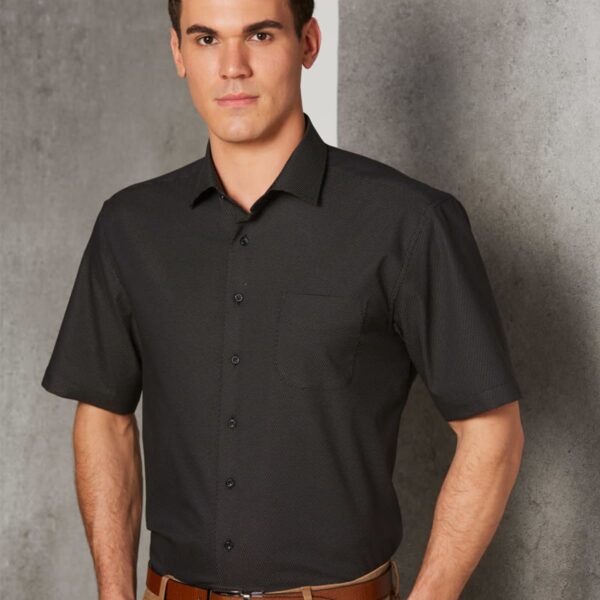 Man wearing a short-sleeve black shirt and khaki pants stands against a gray background.