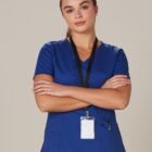 A woman in blue scrubs with a name badge stands with arms crossed, looking at the camera.