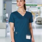 A woman in blue scrubs stands in a hospital hallway with medical equipment behind her.