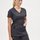Woman in dark scrubs standing with hand in pocket, smiling, against a plain background.