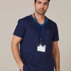 Man in navy blue scrubs with name badge, standing with one hand in his pocket.