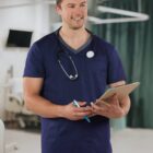 Smiling male nurse in blue scrubs holding a clipboard and pen in a hospital room.