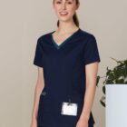 Smiling person in navy blue scrubs with a badge, standing indoors near a plant.