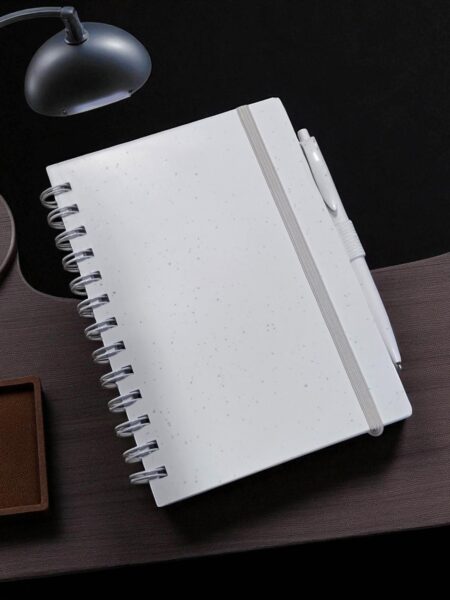 A white spiral notebook for event planning with custom compendiums on a dark desk under a lamp.