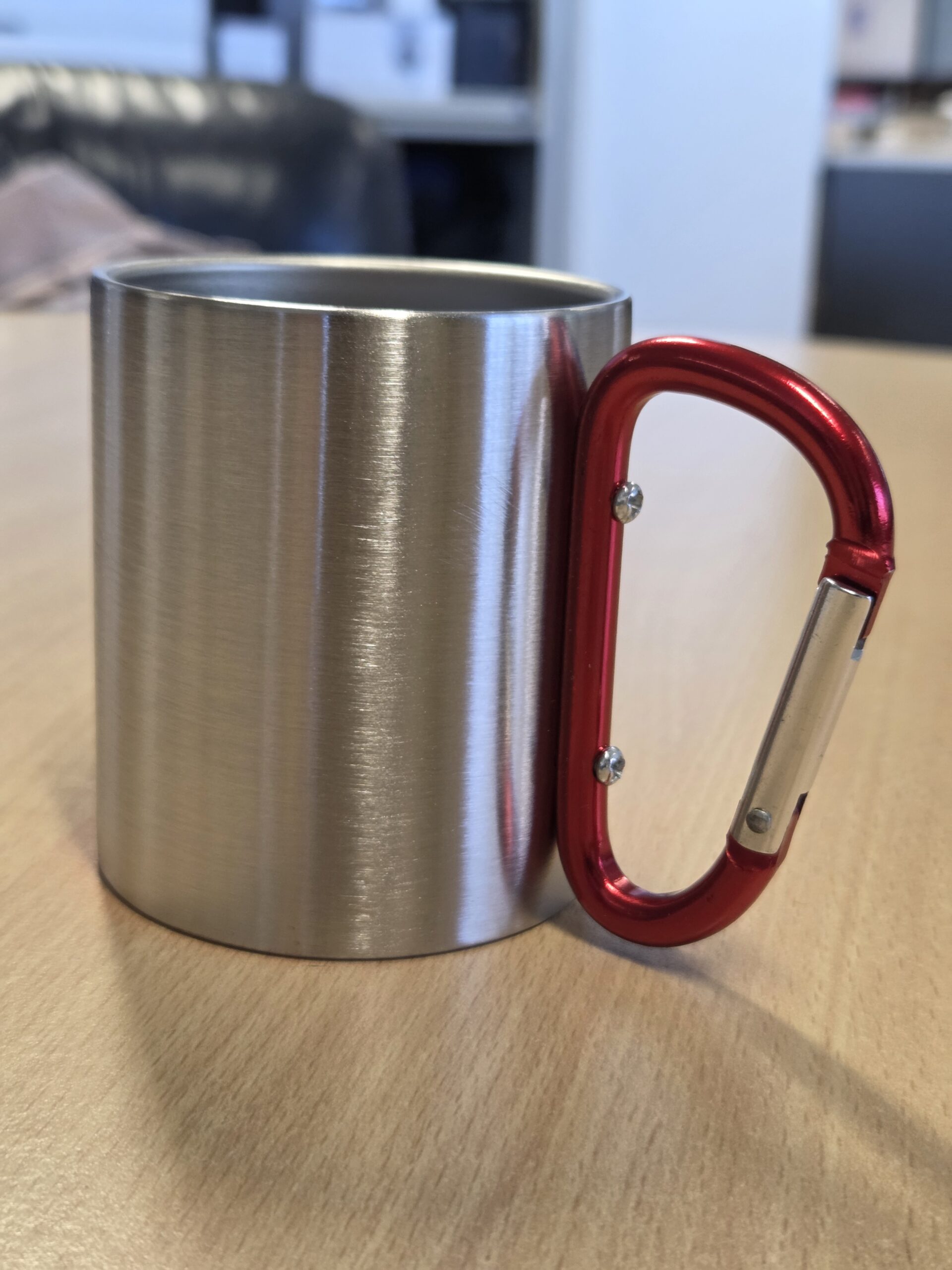 A metal merchandise mug with a red carabiner handle on a wooden table.