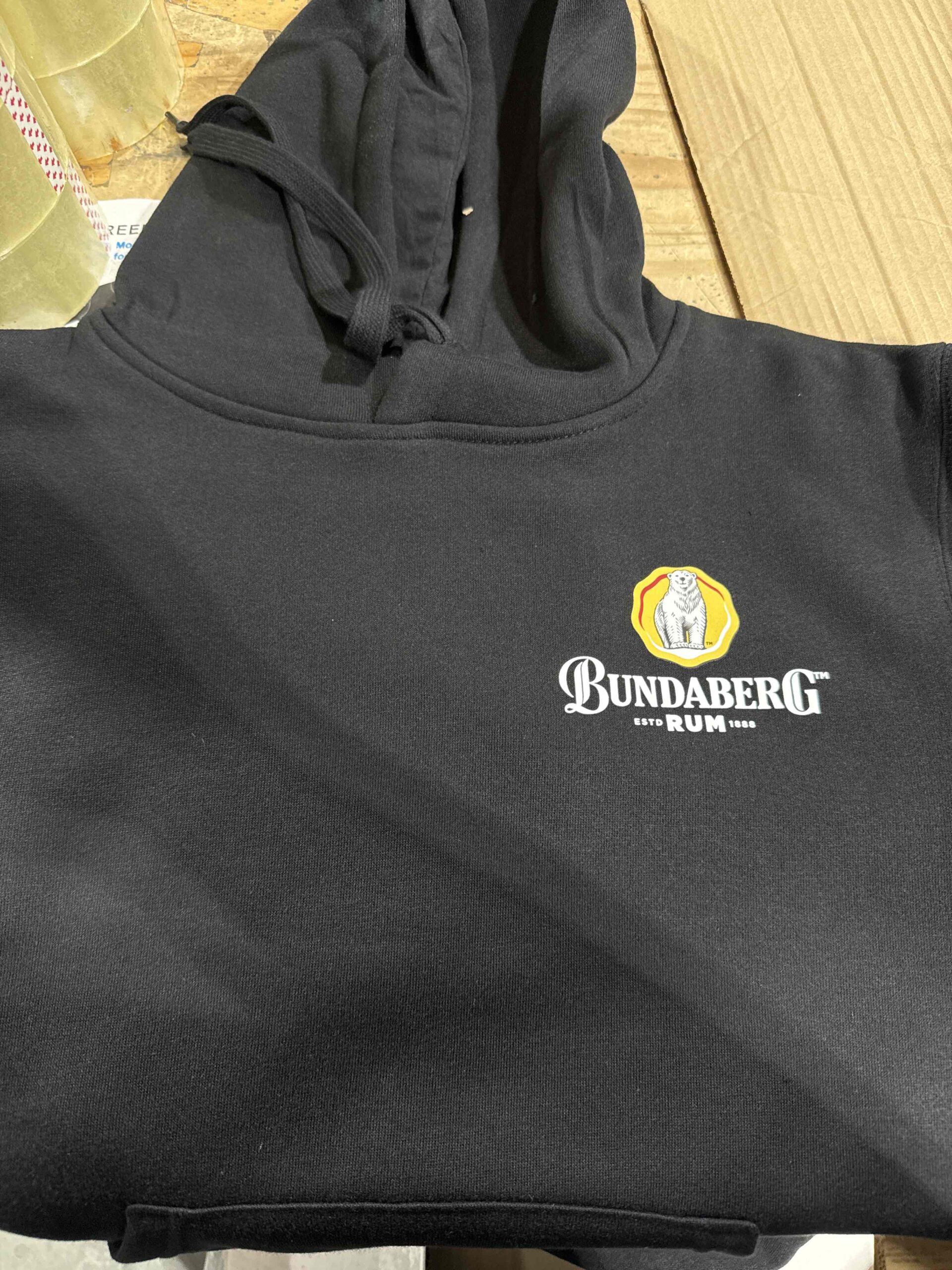 Black Bundaberg Rum hoodie with white logo and yellow bear emblem on a wooden surface.