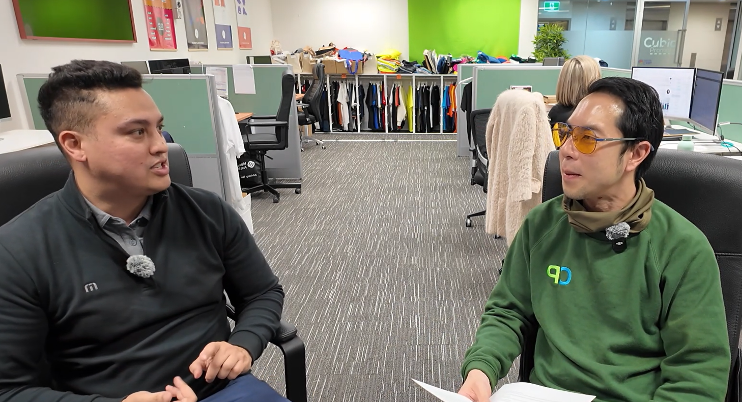 Two men discuss corporate golf in an open-plan office, one holding papers by the desks.