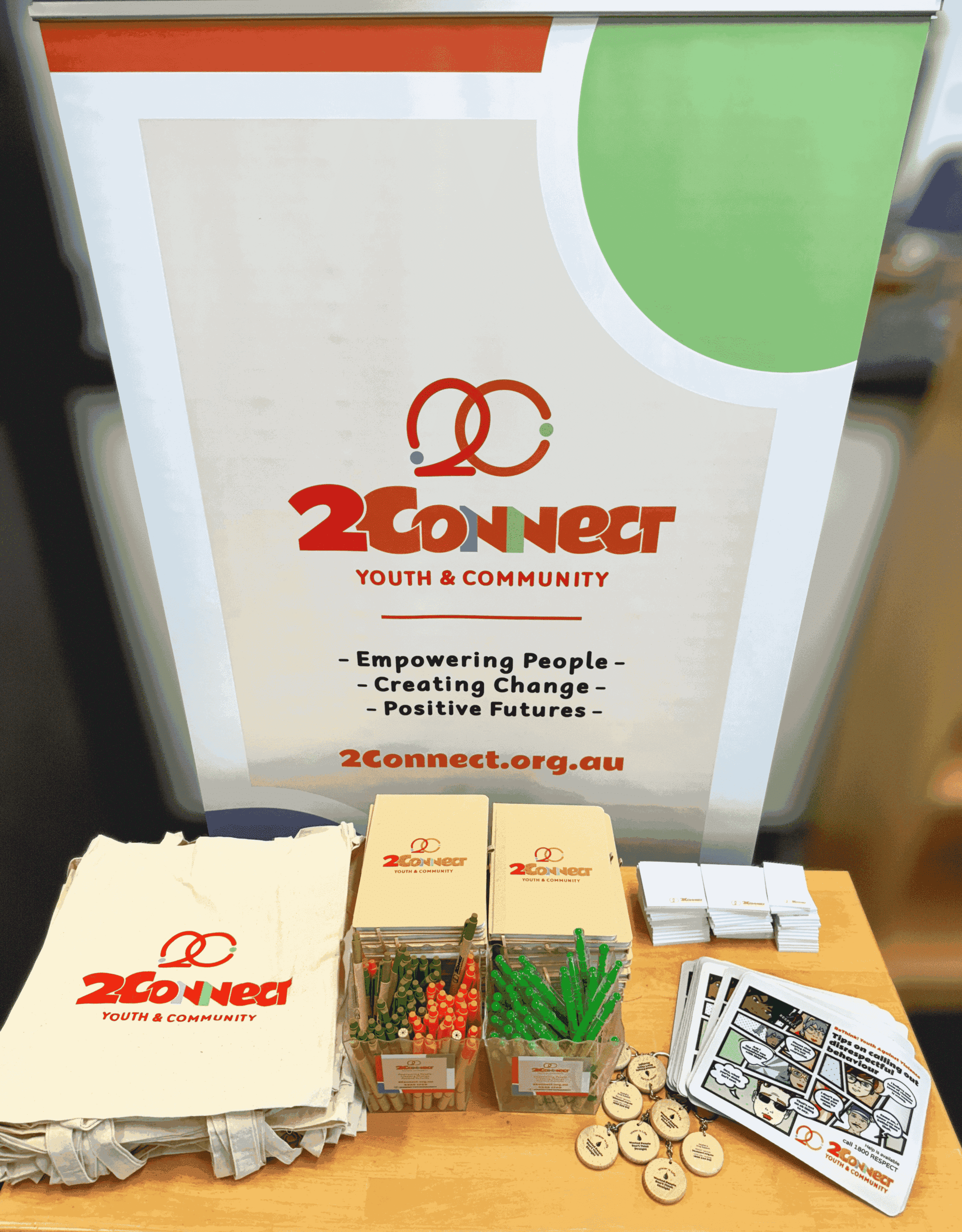 Table with 2Connect Youth & Community merchandise: tote bags, pens, badges, and brochures.