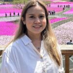 A woman in a white shirt smiles amid vibrant pink flower fields on a sunny day.