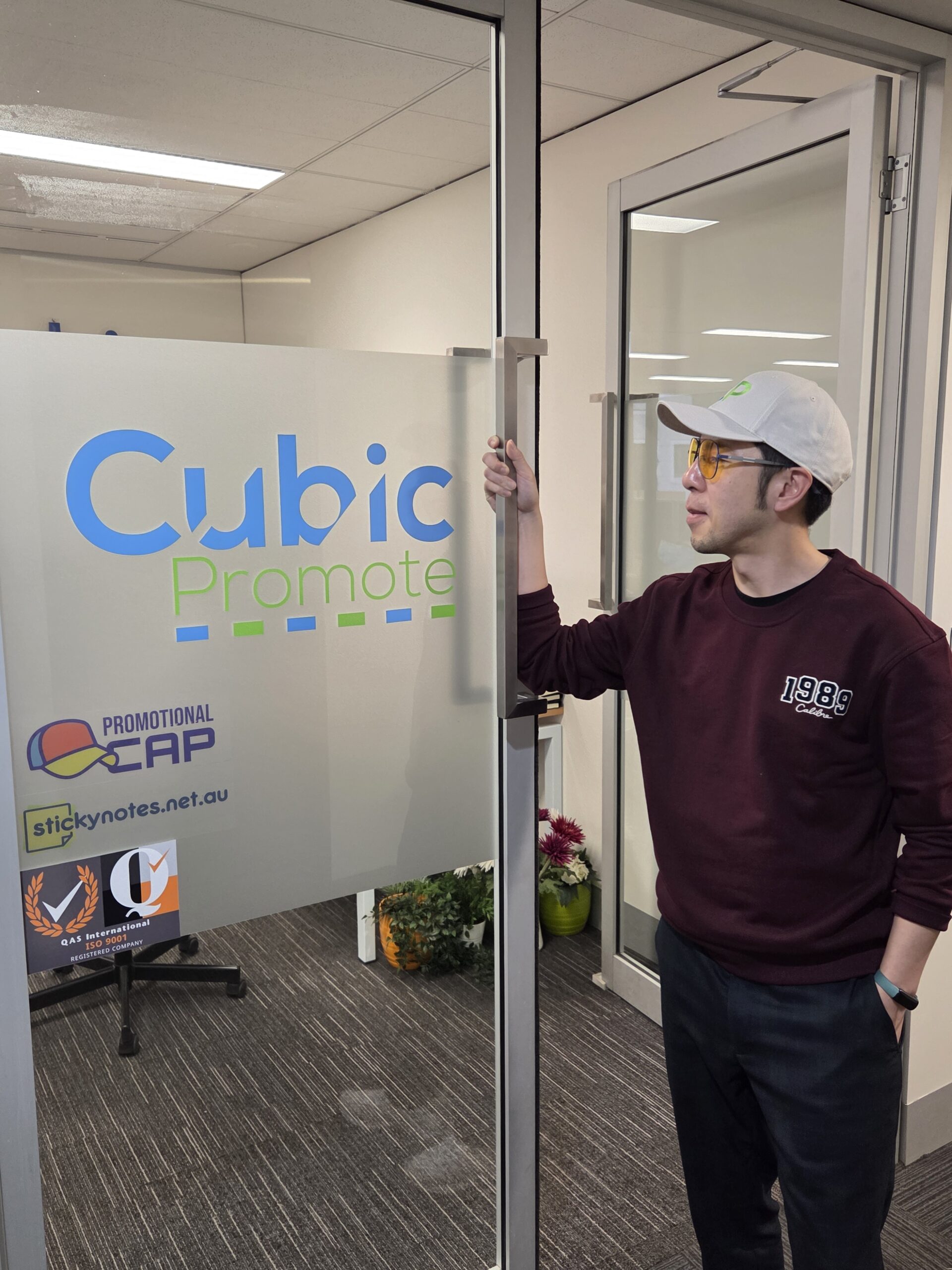 Man in maroon sweater and cap opens office door with Cubic Promote custom merchandise sign.