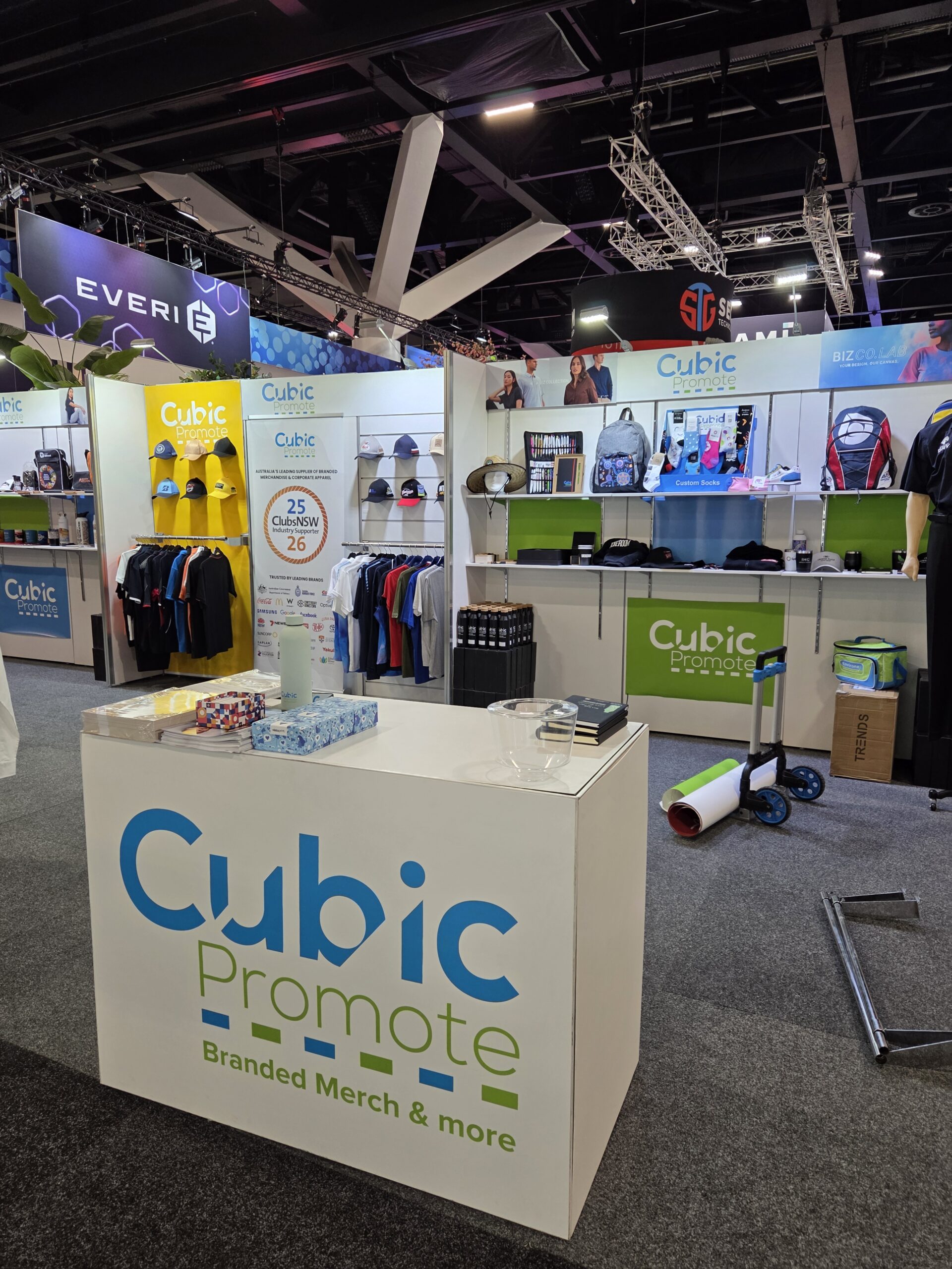 Trade show booth displaying Cubic Promote merch at the Australasian Gaming Expo: shirts, hats, bags.