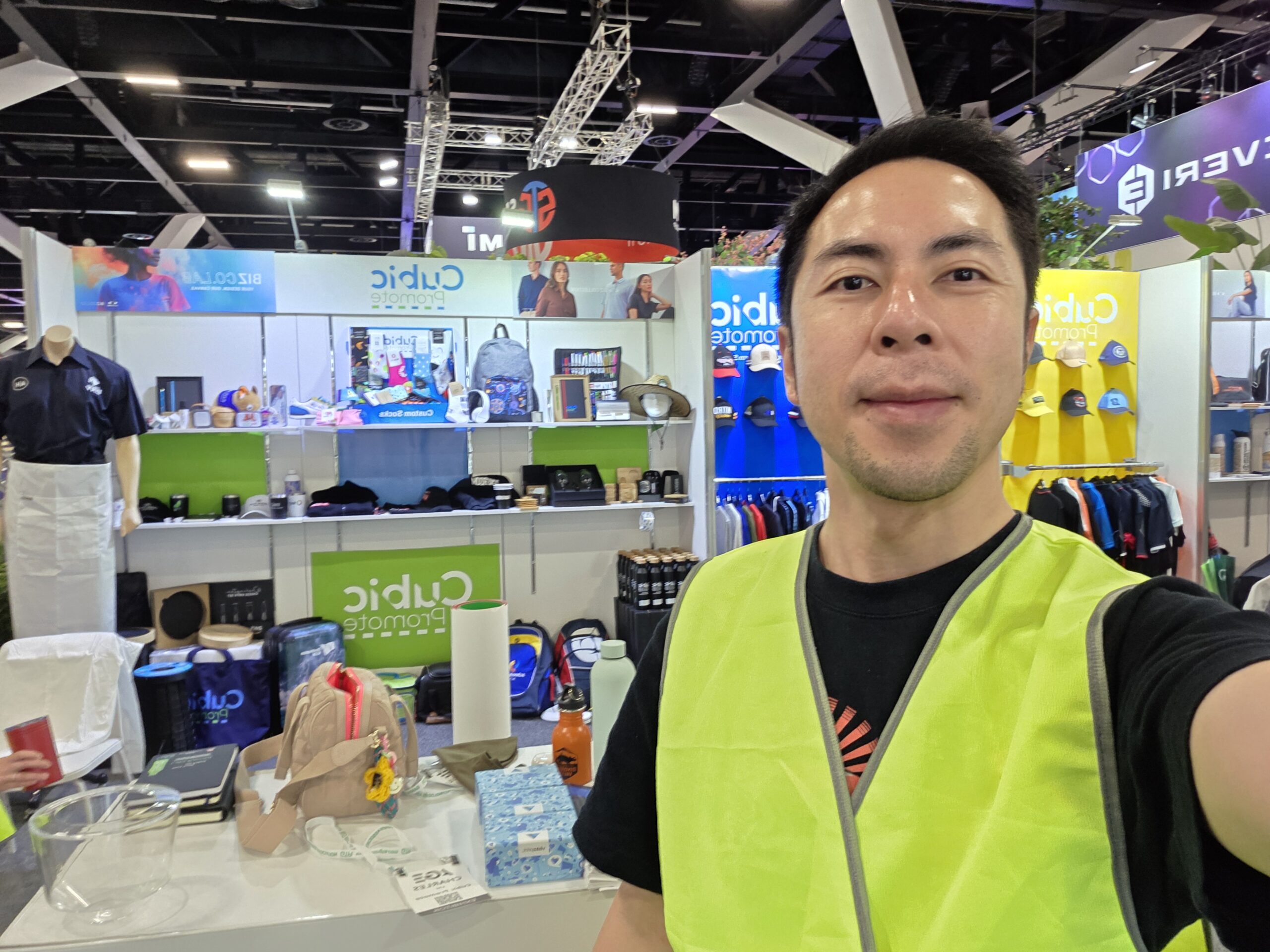 A man in a neon vest smiles at a gaming expo booth with colorful shirts, hats, and displays.