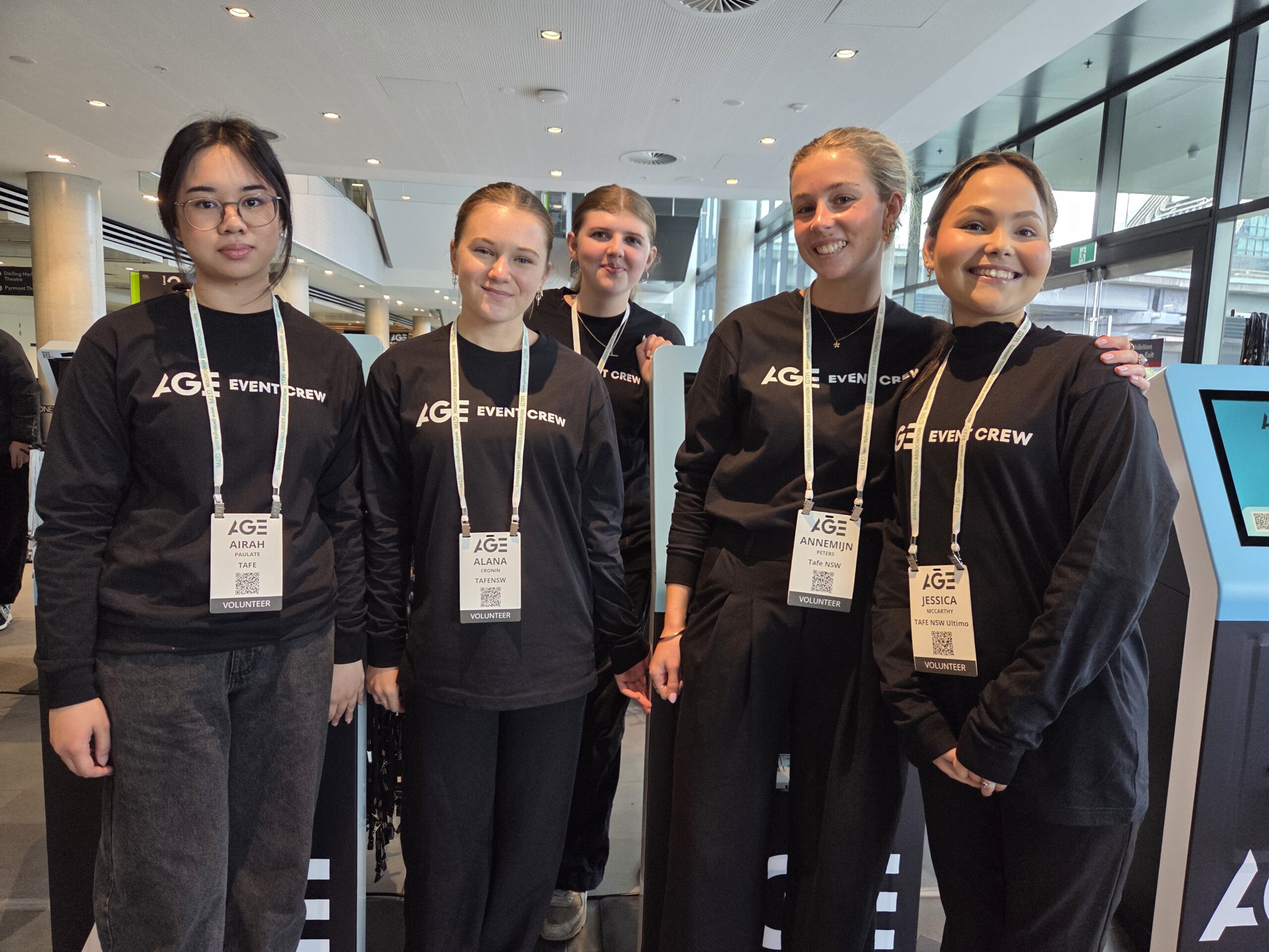 Five women in black AGE EVENT CREW shirts smile indoors at the 2025 Australian Gaming Expo.