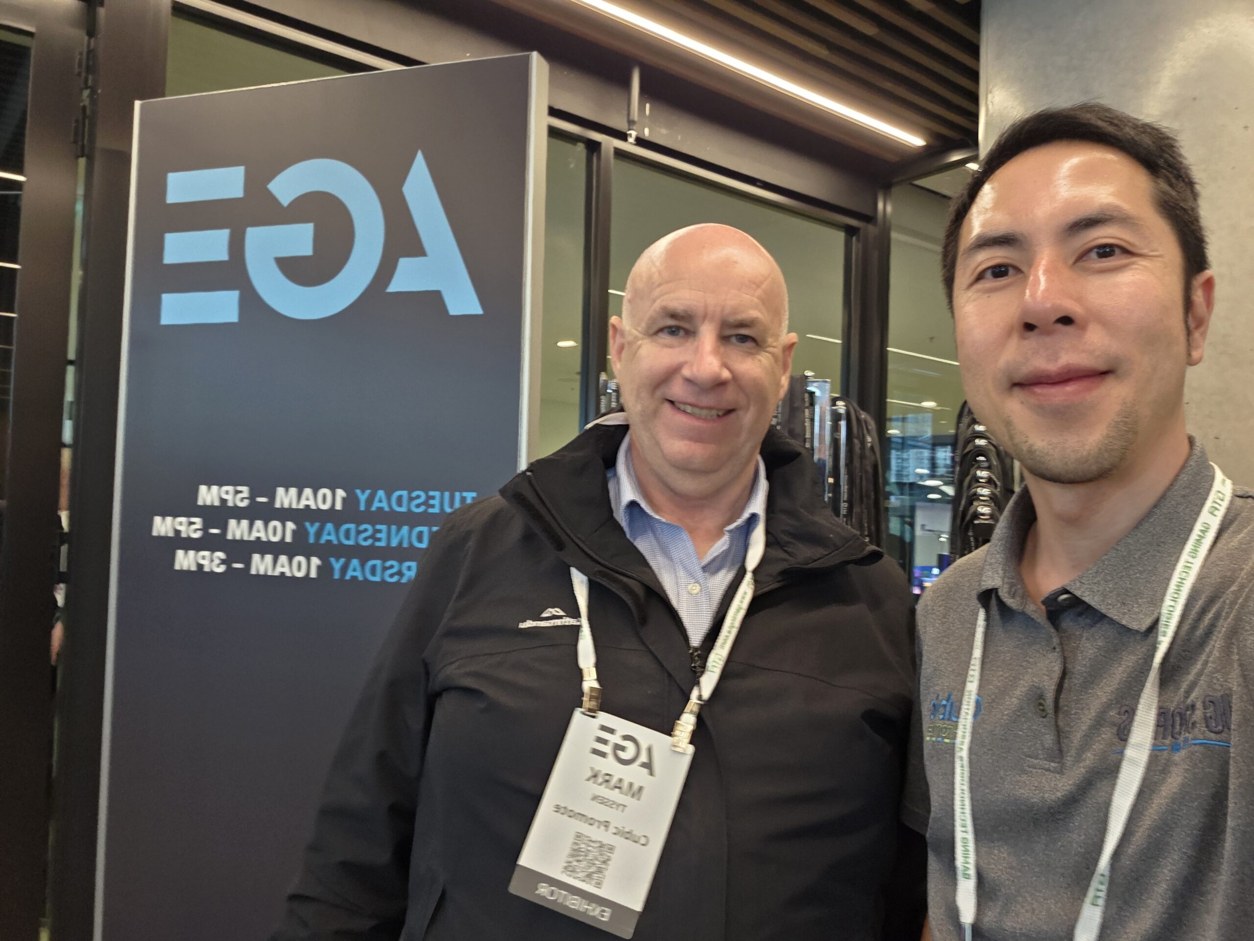 Two men smiling at Day 1 of the 2025 Australian Gaming Expo, AGE event sign in the background.