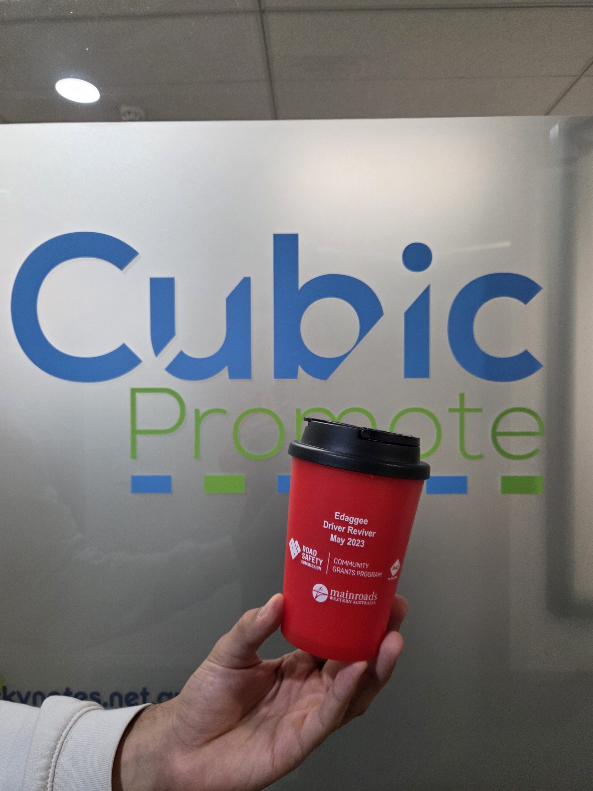 A hand holds a red thermal mug in front of a frosted glass sign reading Cubic Promote.