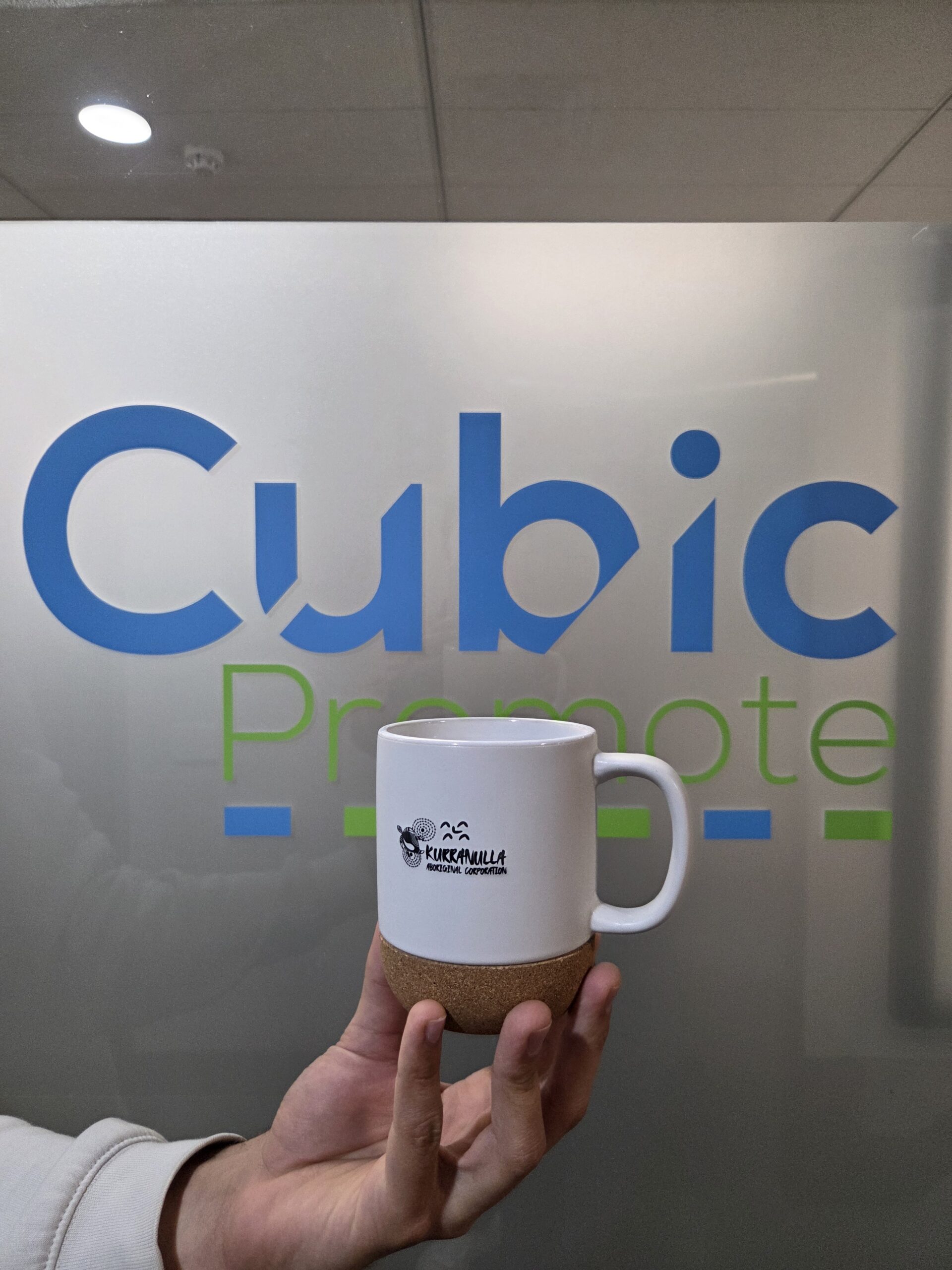 Hand holding a Ceramic Mug with a logo, in front of a frosted glass Cubic Promote sign.