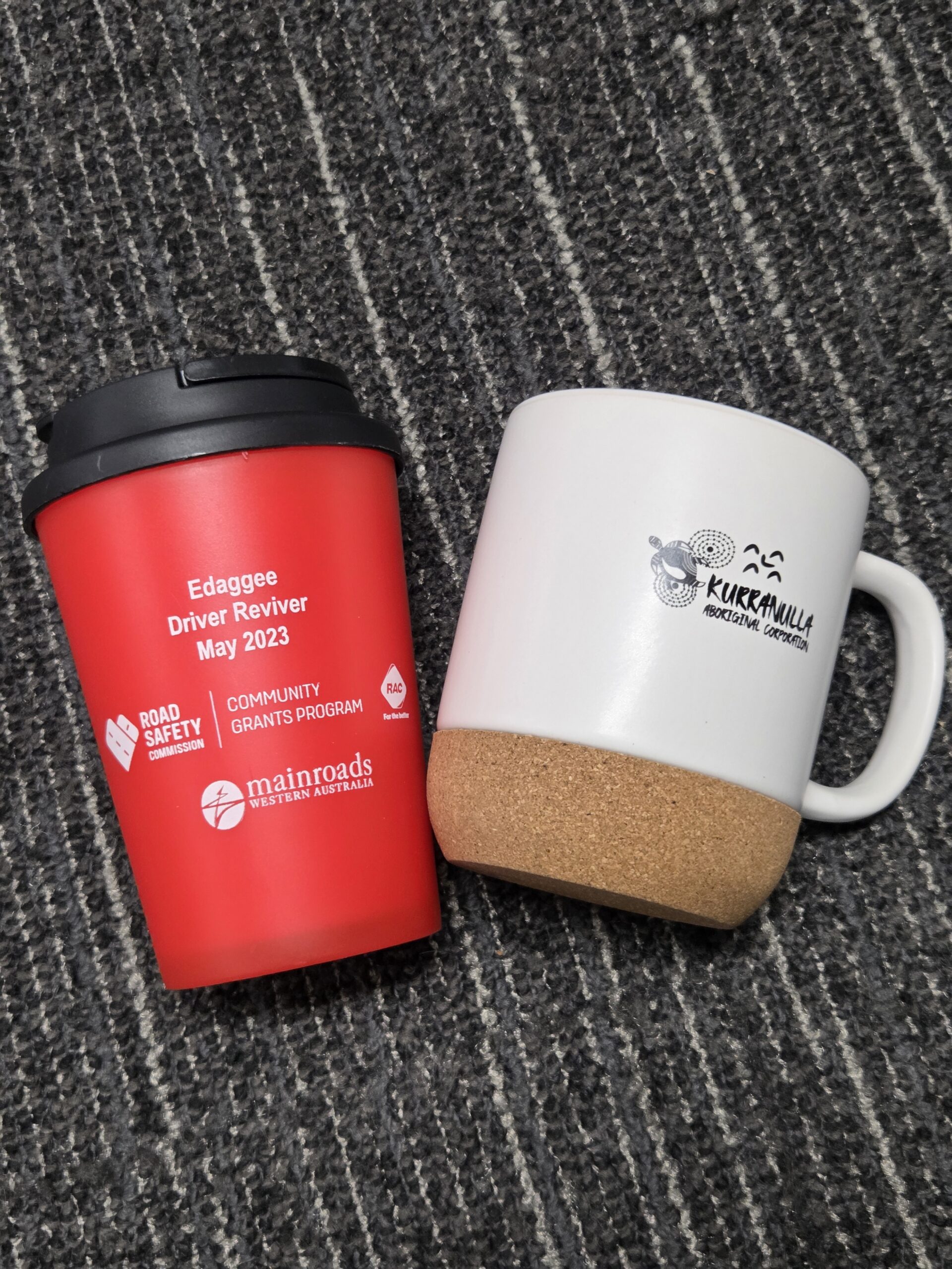 Two reusable thermal mugs, one red and one white, rest on a dark striped carpet.