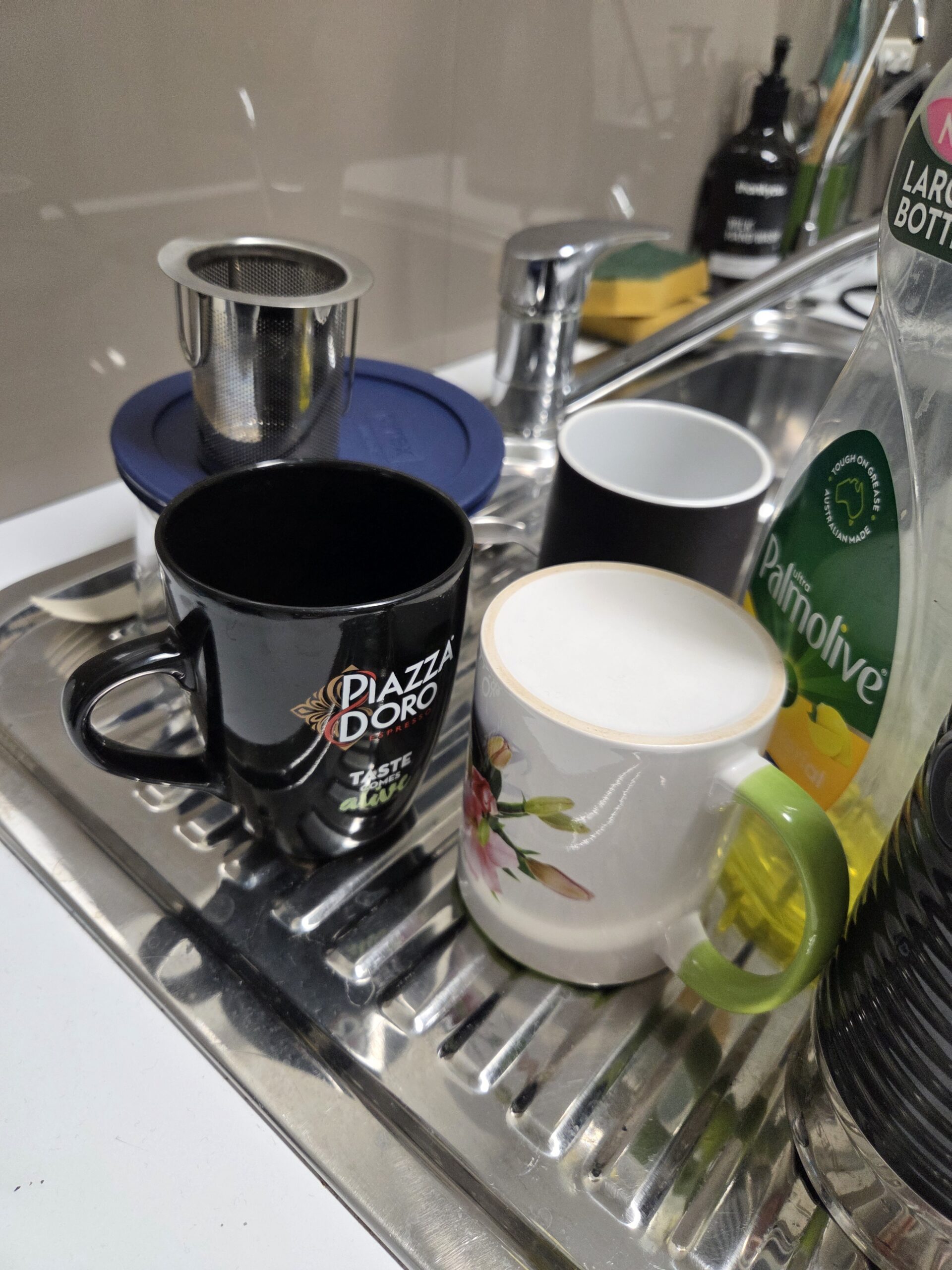 Three ceramic mugs and a tea strainer rest on a dish rack by the sink with soap and a sponge.