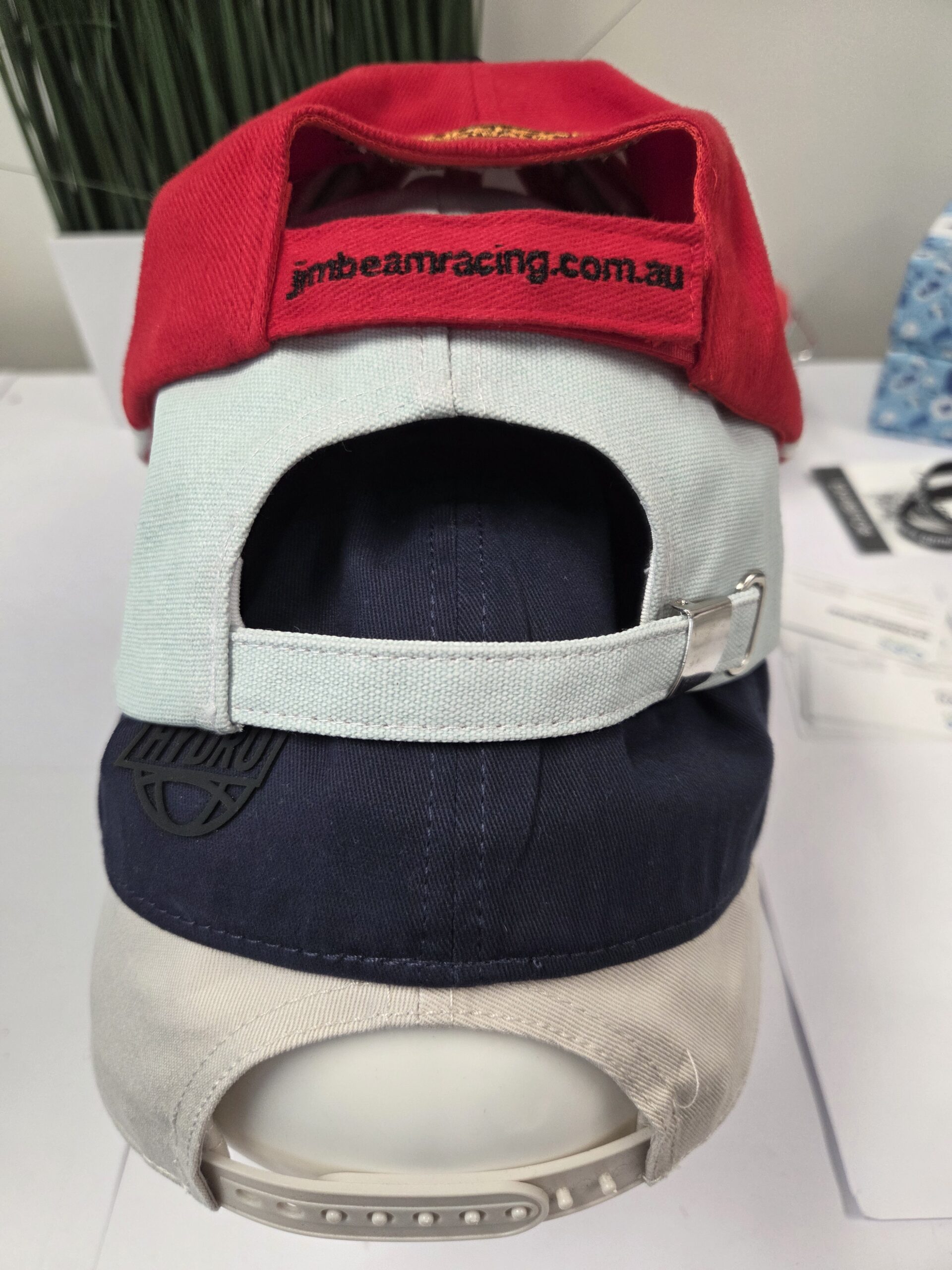 Three branded caps stacked, each showing cap closures and back straps, on a white surface.