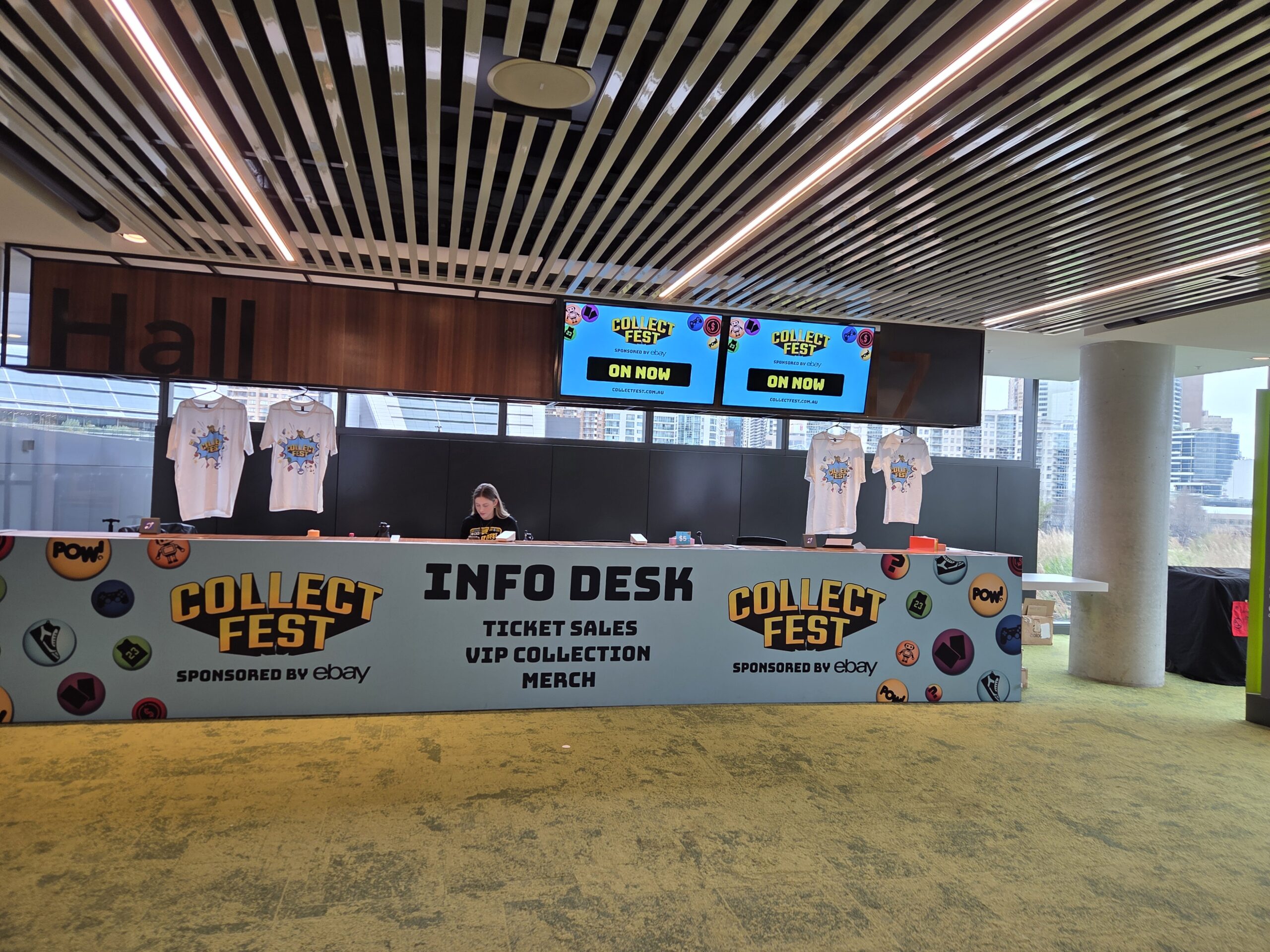A Collectfest Sydney 2025 info desk with custom-branded merchandise and a staff member behind the counter.