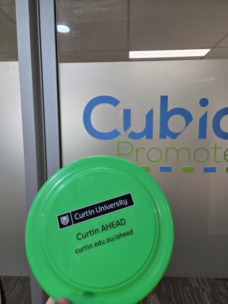 A green promotional frisbee from Curtin University held in front of a glass door with Cubico Promote text.