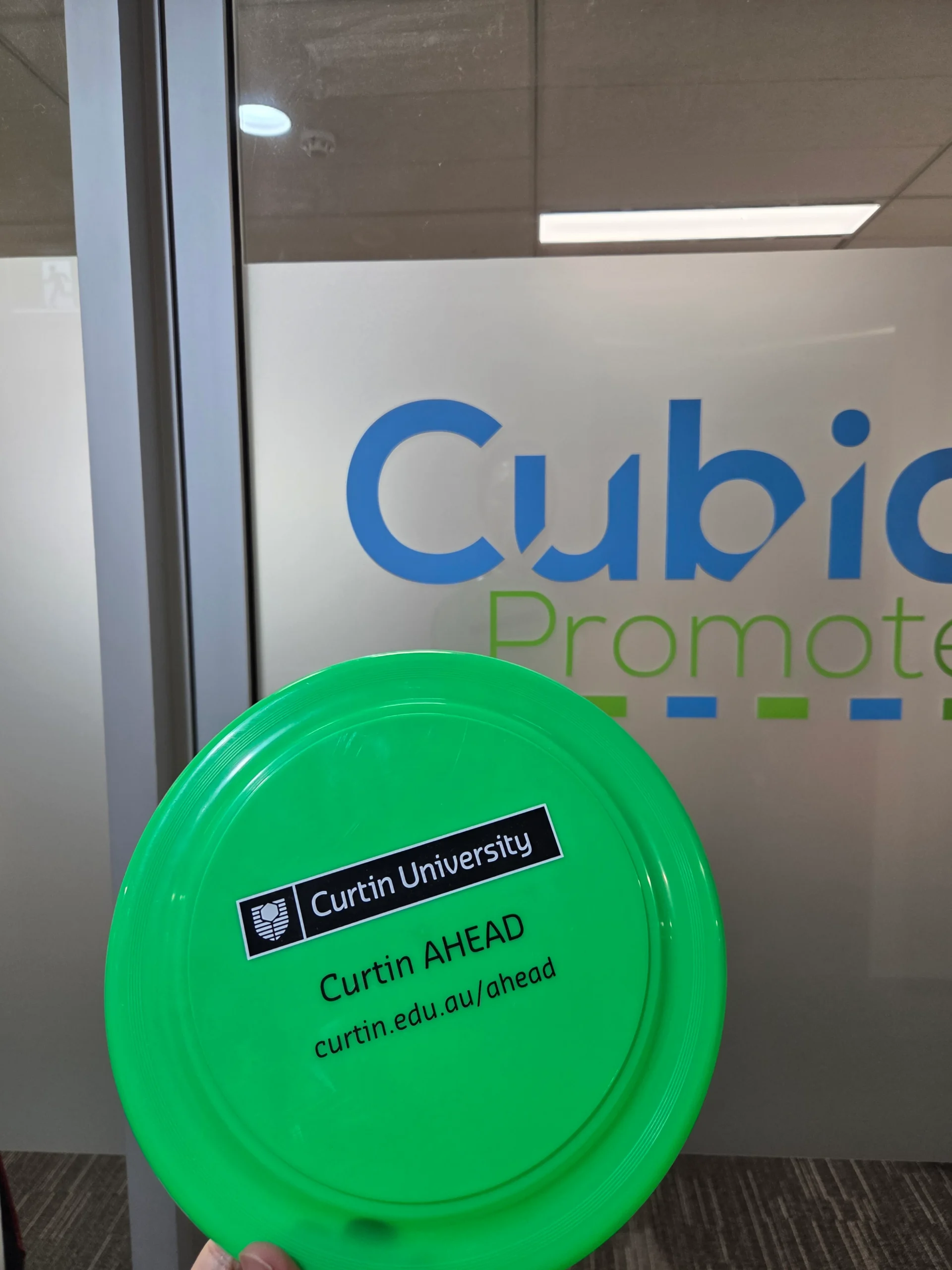 A green frisbee from Curtin AHEAD showcased among other frisbees by a glass office door.