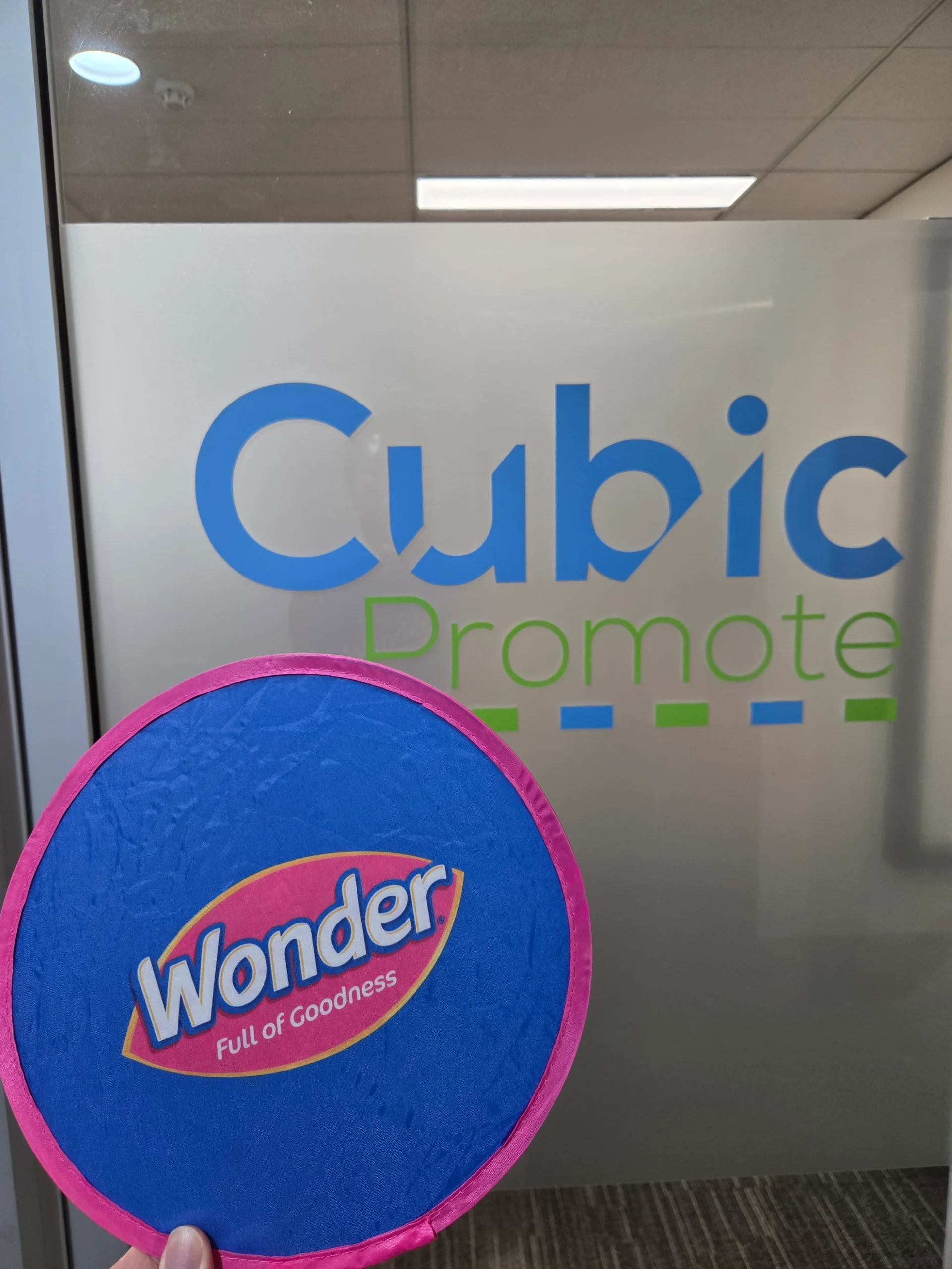 Hand holding one of our frisbees in front of a blue and pink Cubic Promote office sign.