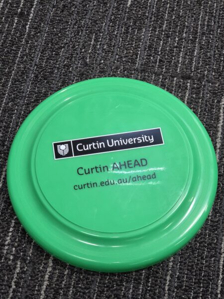 A green Promotional Frisbee with Curtin University and Curtin AHEAD logos sits on a carpet.
