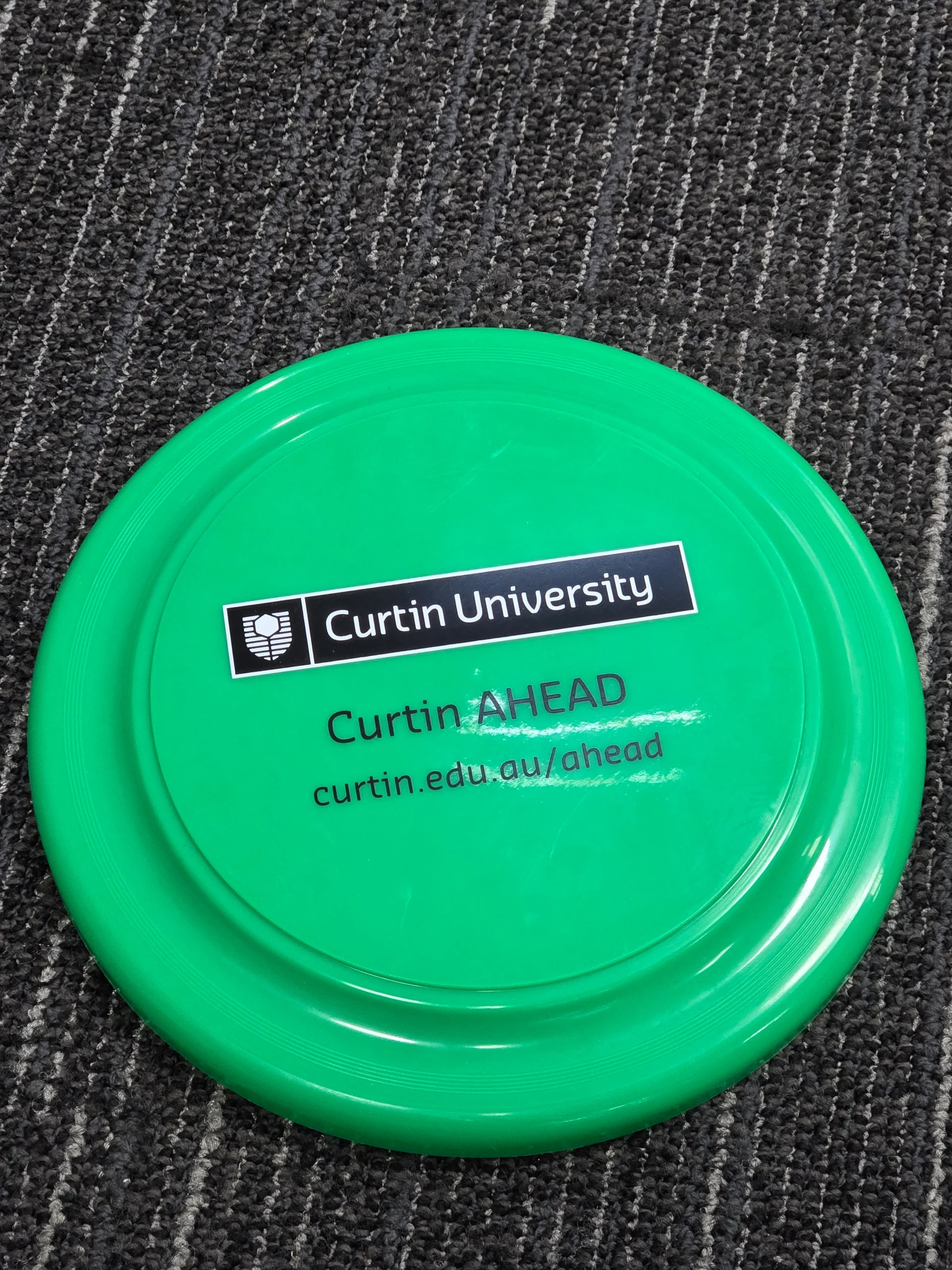 Frisbees with Curtin University Curtin AHEAD text on a green disc atop a patterned carpet.