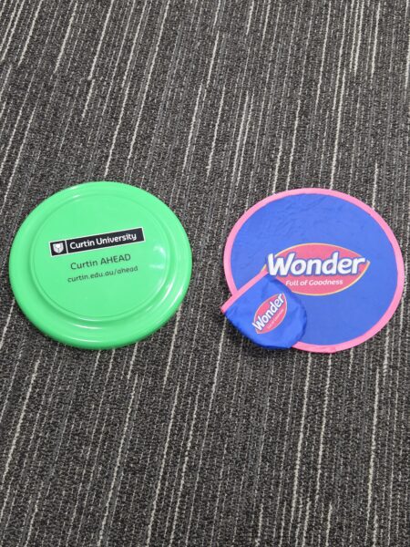 A green Promotional Frisbee and a blue Wonder paddle with a ball on a striped carpet.