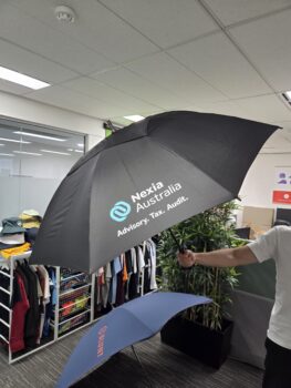 Person holding a black Nexia Australia umbrella in an office with umbrellas, clothes, and plants nearby.