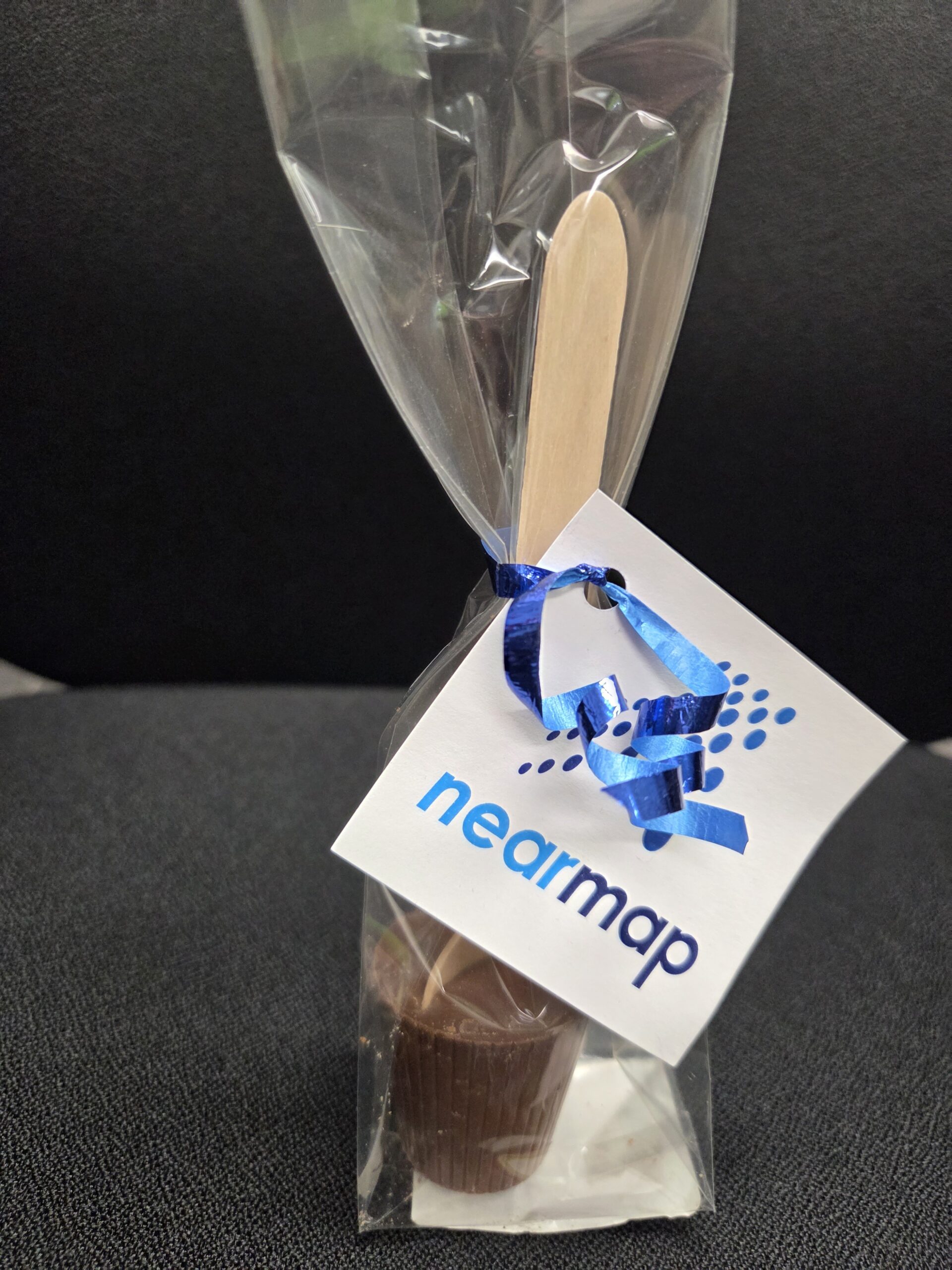 A chocolate-coated wooden stick lollipop, wrapped in plastic with a blue ribbon and Nearmap tag.
