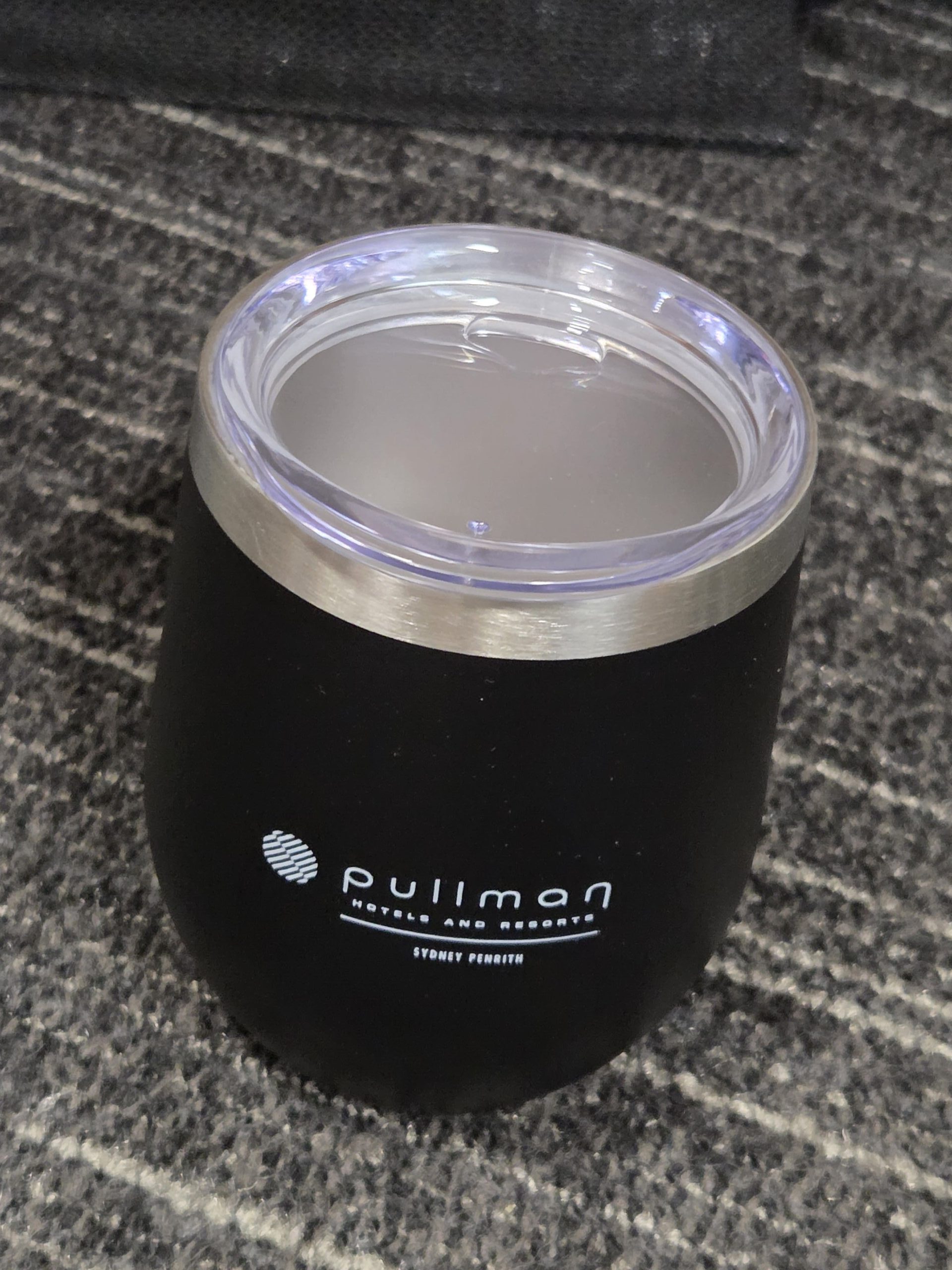 Black Pullman-branded tumbler with clear lid at Pullman Hotels Sydney on a gray textured carpet.