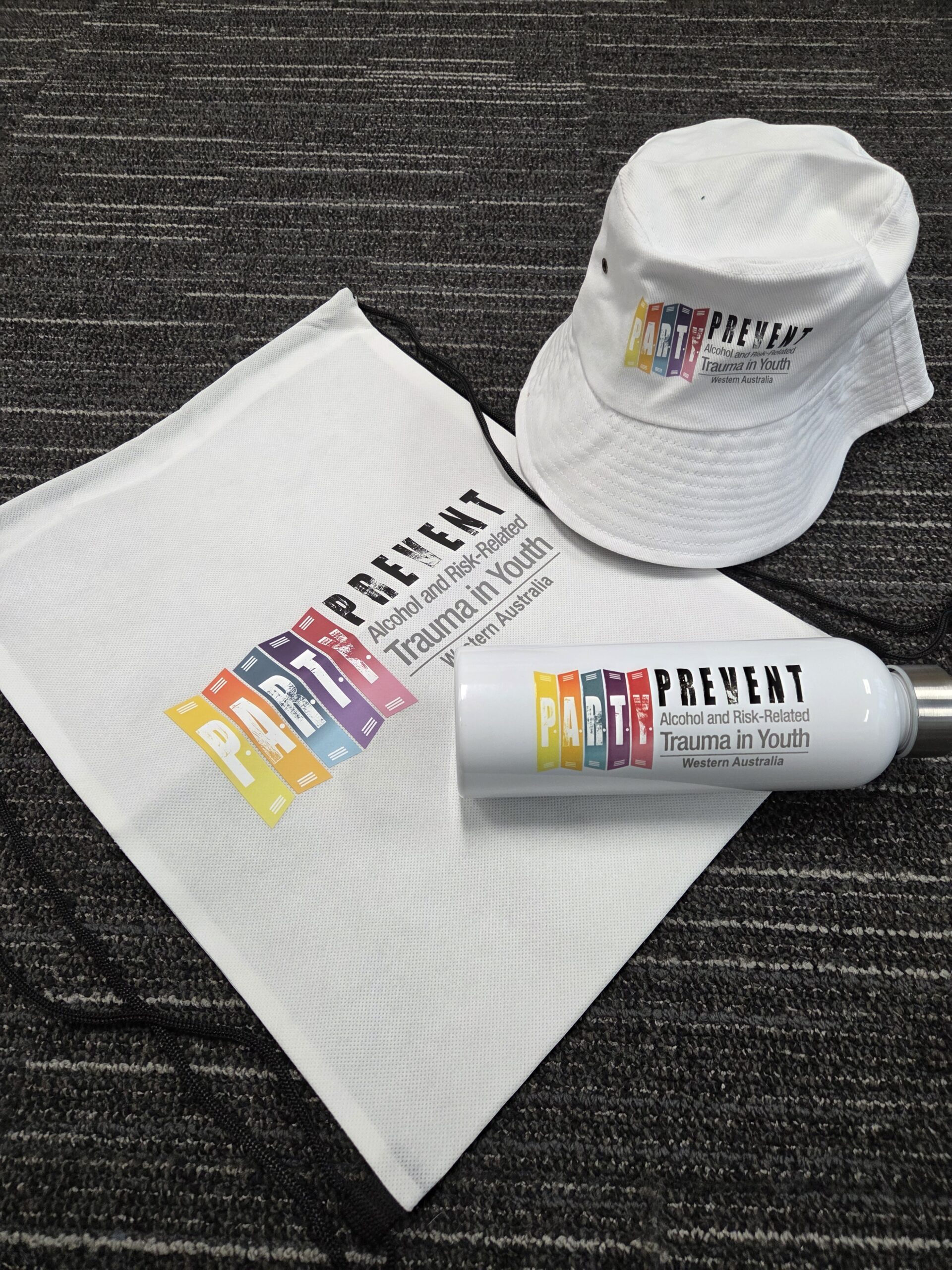 White hat, tote bag, and drink bottle with colorful Royal Perth Hospital youth trauma logo.