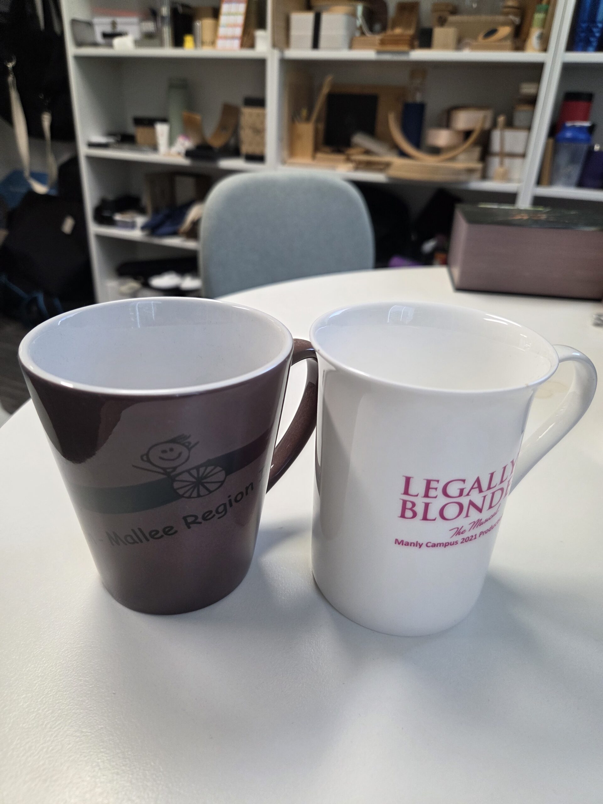 Two promotional mugs on a white table with shelves of items in the background.