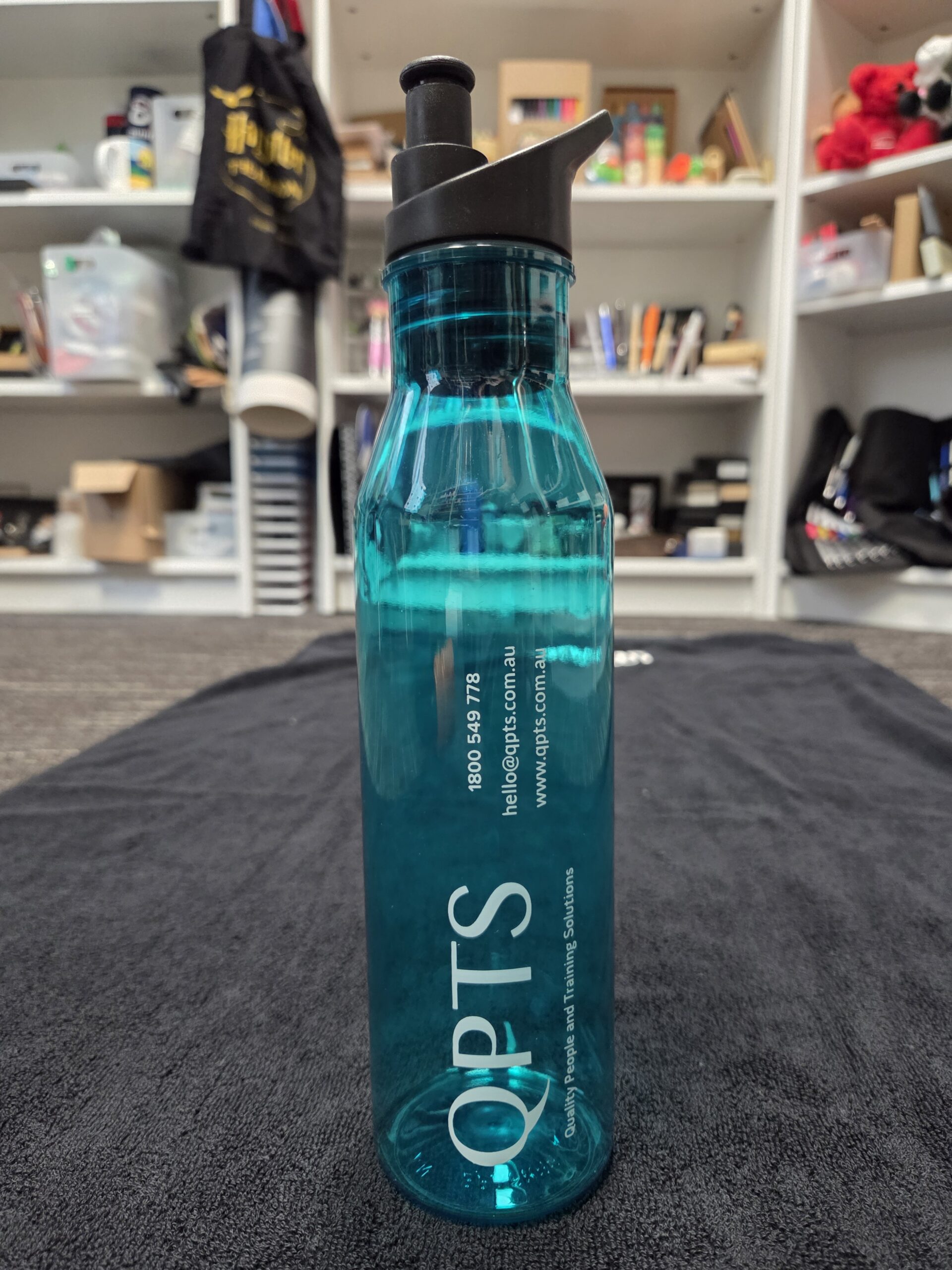 A teal OPTS water bottleāwondering which bottle?ārests on a black towel with shelves behind.