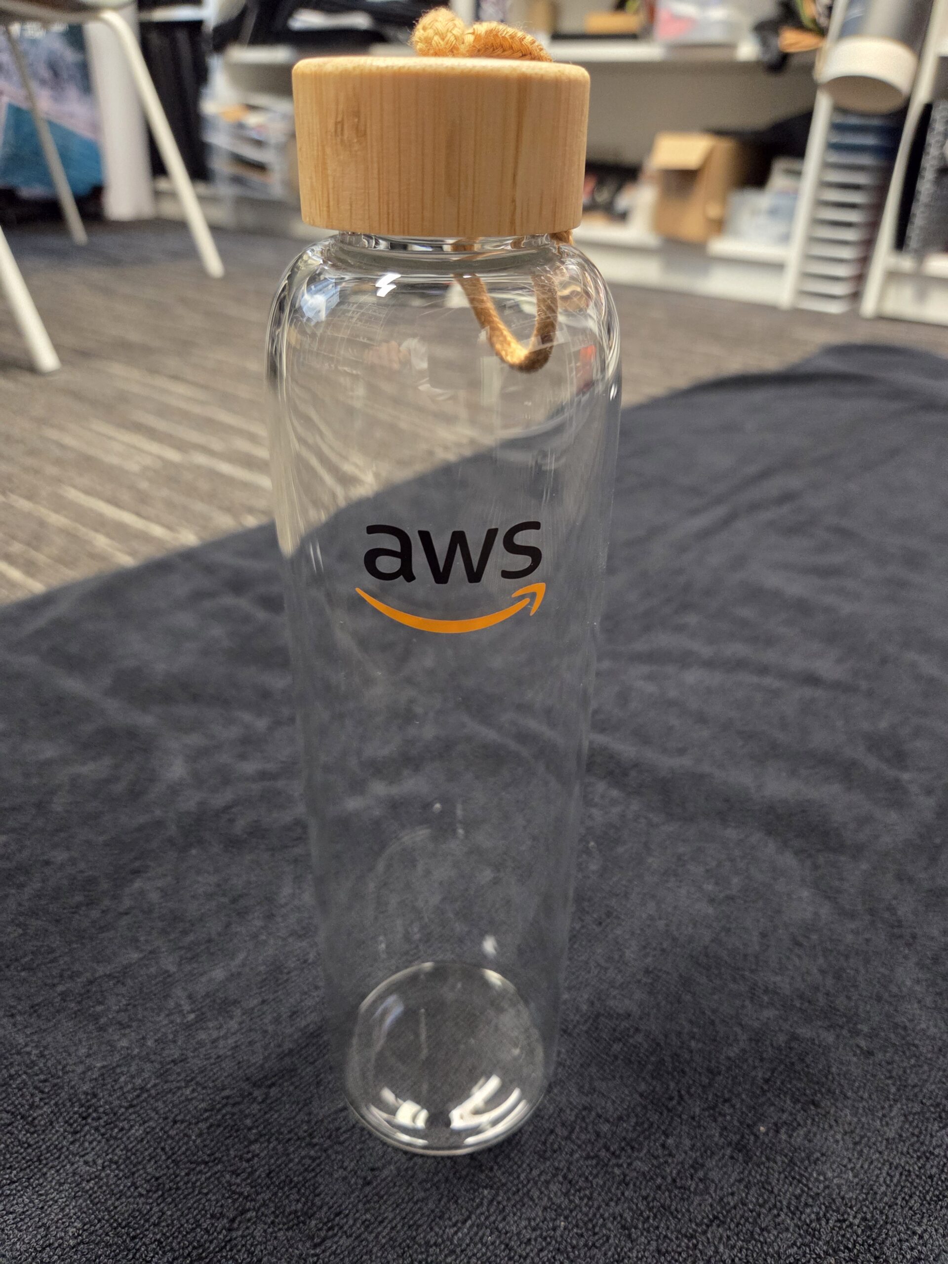 AWS Branded Glass Drink Bottle with bamboo lid, set on a dark cloth surface.